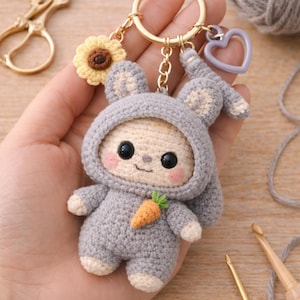 May include: A handmade crochet bunny keychain in gray with a small orange carrot. The bunny has a hood and is attached to a gold keyring with a sunflower and a purple heart charm. The keychain is approximately 3 inches tall.