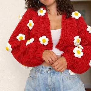 Peut inclure: Un cardigan rouge en crochet, orné de marguerites blanches et jaunes. Ce modèle ouvert et court présente des manches longues. Le cardigan en tricot épais est porté avec un jean en denim bleu clair. Mesure environ 50 cm de long.