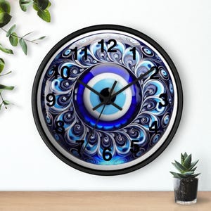 Puede incluir: Un reloj de pared redondo con marco negro. La esfera presenta un diseño de ojo azul y blanco con números y manecillas negras. El reloj está sobre una superficie de madera con una pequeña planta en maceta.
