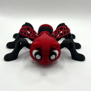 Puede incluir: Un juguete de insecto rojo y negro impreso en 3D, con grandes ojos blancos y una expresión sonriente. Tiene seis patas negras y dos alas rojas estampadas. El diseño es caricaturesco y caprichoso.