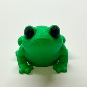 Pode incluir: Uma estatueta de sapo verde brilhante com olhos grandes e pretos. O sapo &eacute; impresso em 3D e tem uma superf&iacute;cie texturizada. O sapo est&aacute; sobre uma superf&iacute;cie branca. Este sapo decorativo &eacute; uma adi&ccedil;&atilde;o divertida a qualquer cole&ccedil;&atilde;o.