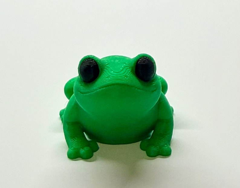 Pode incluir: Uma estatueta de sapo verde brilhante com olhos grandes e pretos. O sapo &eacute; impresso em 3D e tem uma superf&iacute;cie texturizada. O sapo est&aacute; sobre uma superf&iacute;cie branca. Este sapo decorativo &eacute; uma adi&ccedil;&atilde;o divertida a qualquer cole&ccedil;&atilde;o.