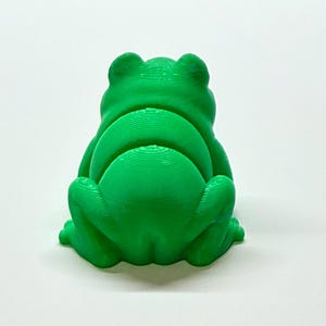 Pode incluir: Uma estatueta de sapo verde brilhante, vista por tr&aacute;s, com uma superf&iacute;cie texturizada. O sapo est&aacute; sentado com as pernas dobradas e as costas arqueadas. A estatueta &eacute; feita de um material s&oacute;lido e est&aacute; sobre um fundo branco.