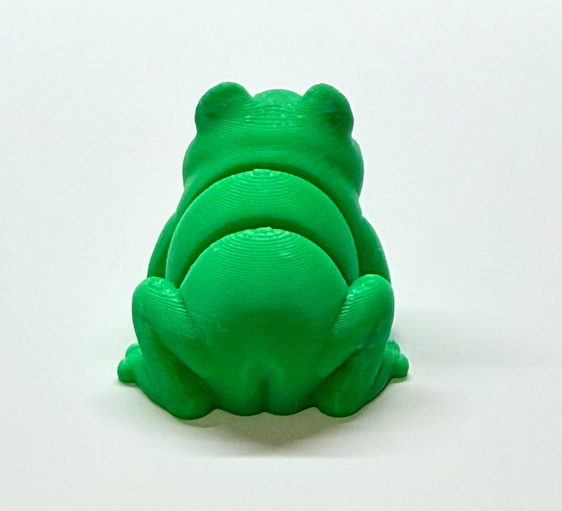 Pode incluir: Uma estatueta de sapo verde brilhante, vista por tr&aacute;s, com uma superf&iacute;cie texturizada. O sapo est&aacute; sentado com as pernas dobradas e as costas arqueadas. A estatueta &eacute; feita de um material s&oacute;lido e est&aacute; sobre um fundo branco.