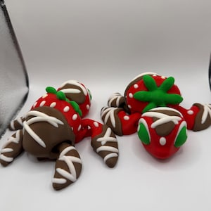 Può includere: Due figure scolpite a forma di granchio con corpi rossi a pois bianchi e cime verdi. Gli arti sono marroni con strisce bianche simili a glassa, che ricordano fragole ricoperte di cioccolato. Ogni figura ha una consistenza liscia e argillosa.