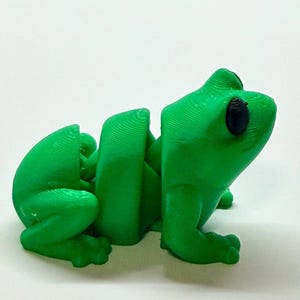 Pode incluir: Uma estatueta de sapo verde brilhante, impressa em 3D, com partes do corpo segmentadas. O sapo tem olhos pretos grandes e est&aacute; sentado. A estatueta &eacute; feita de pl&aacute;stico e est&aacute; sobre uma superf&iacute;cie branca.