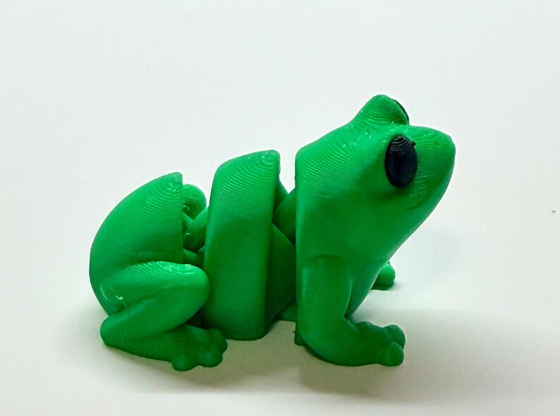 Pode incluir: Uma estatueta de sapo verde brilhante, impressa em 3D, com partes do corpo segmentadas. O sapo tem olhos pretos grandes e est&aacute; sentado. A estatueta &eacute; feita de pl&aacute;stico e est&aacute; sobre uma superf&iacute;cie branca.