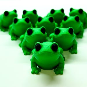 Pode incluir: Um grupo de figuras de sapos verdes com olhos pretos dispostos em forma&ccedil;&atilde;o triangular. Os sapos s&atilde;o de cor verde vibrante e t&ecirc;m uma textura suave. O fundo &eacute; branco liso.