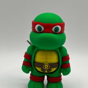 Puede incluir: Una figura verde de una Tortuga Ninja, con una máscara y detalles rojos. La figura tiene un pecho amarillo con un cinturón marrón y la letra "R". Los ojos son blancos con pupilas verdes. La figura está de pie.