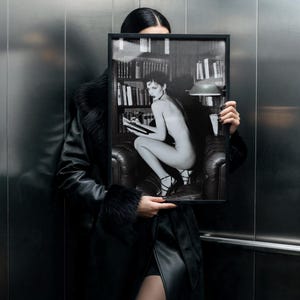 Może przedstawiać: Czarno-biała oprawiona fotografia trzymana przed osobą. Na fotografii widnieje naga osoba siedząca w sk&oacute;rzanym fotelu, czytająca książkę w otoczeniu biblioteki. Osoba ma na sobie czarny sk&oacute;rzany płaszcz.