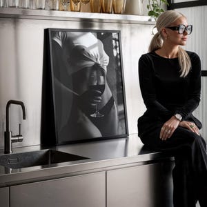May include: A monochrome framed print depicting a person holding a glass of red wine. The print is on a stainless steel surface. A woman in black attire and glasses is seated on the countertop.