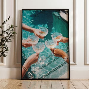 May include: A framed print depicts a celebratory moment. Several hands hold up ornate champagne glasses filled with a golden liquid, set against a backdrop of a turquoise pool. The image evokes a sense of luxury and festivity.
