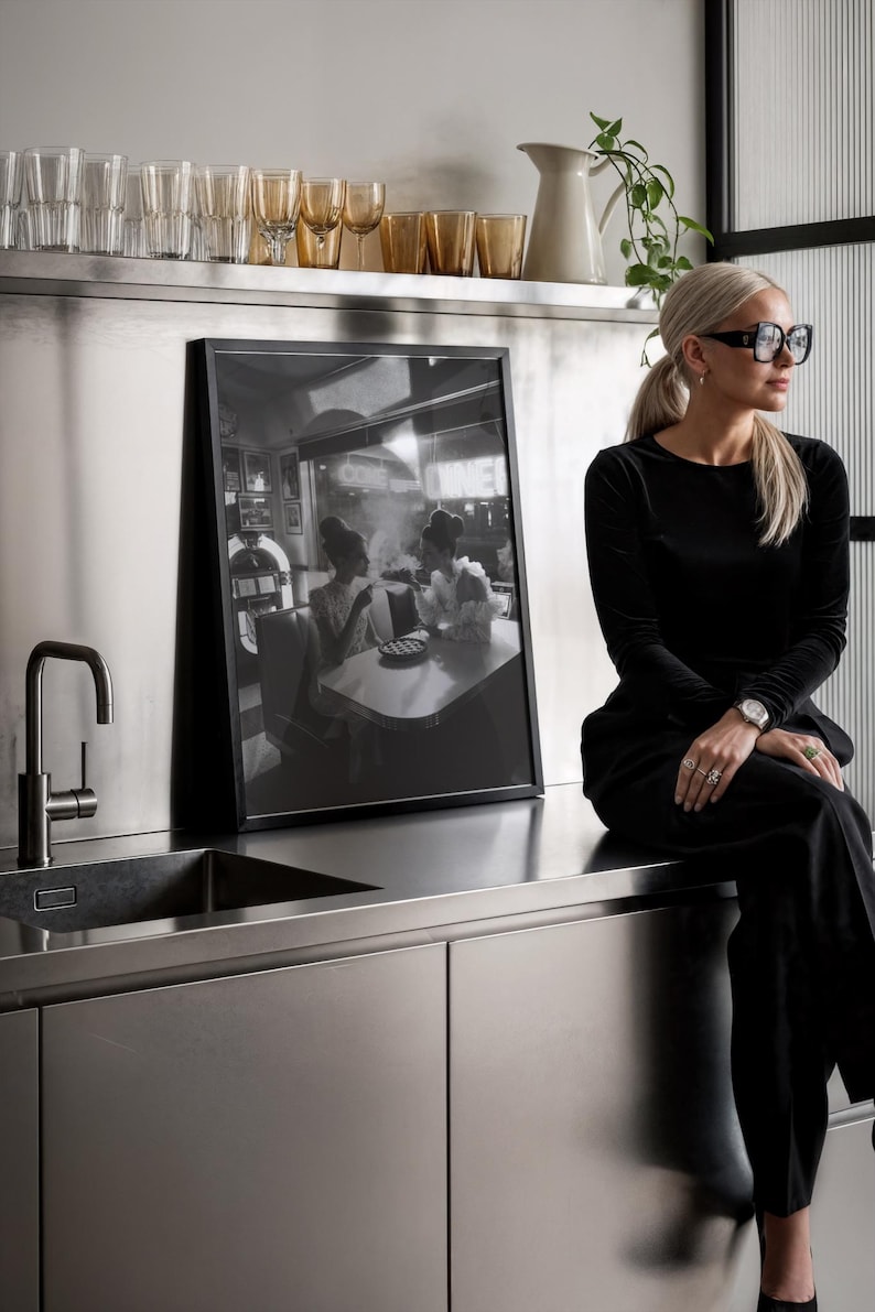 Retro diner ladies poster displayed in modern stainless steel kitchen with woman in sunglasses gold glassware and contemporary design showing black white photography