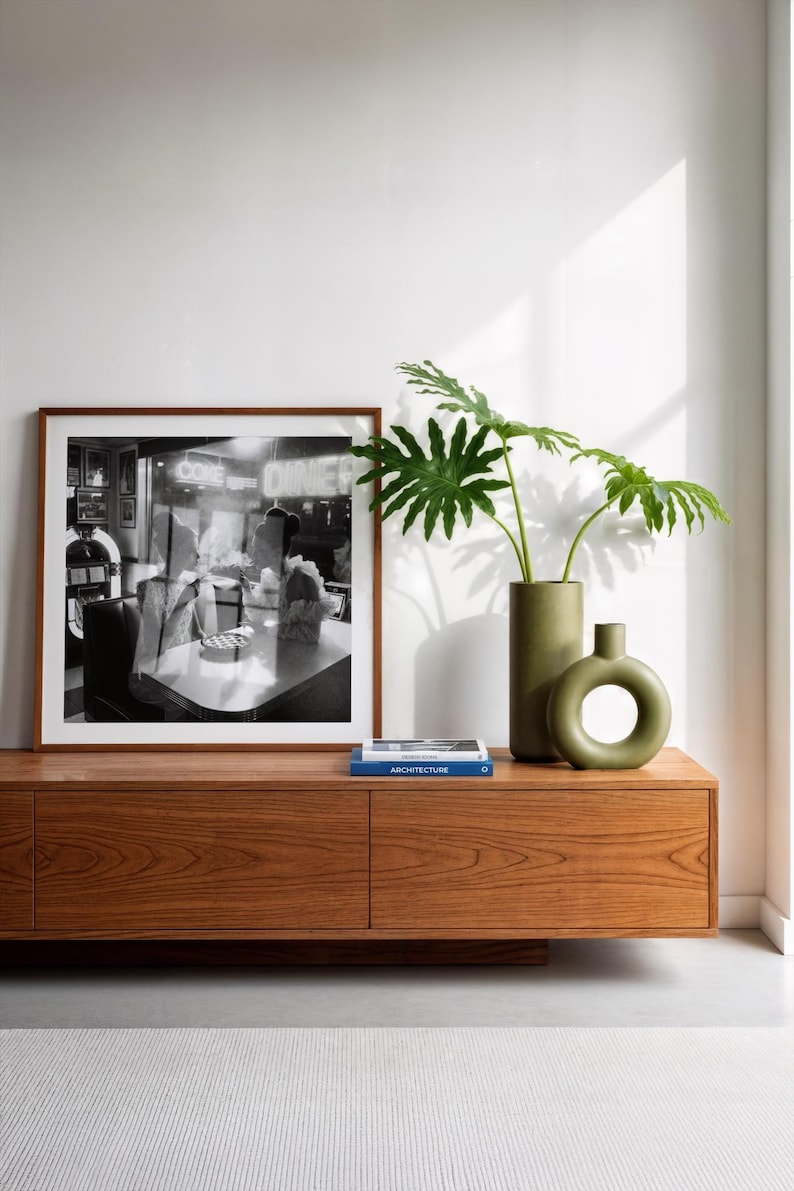 Vintage women diner photo poster in wood frame displayed on mid-century walnut credenza with green ceramic vases tropical plants and architecture books contemporary interior