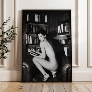 A framed black and white editorial photograph of a sensual woman with dark hair in a classic library setting, kneeling on a leather tufted chair. The striking monochrome print is displayed in a sleek black frame in a modern space.