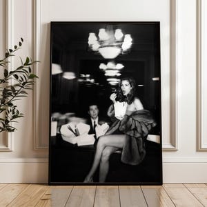 Può includere: Stampa incorniciata in bianco e nero raffigurante una donna che mangia popcorn, avvolta in una pelliccia, in un ristorante. Un uomo in giacca e cravatta è visibile sullo sfondo. La stampa è in una cornice nera.
