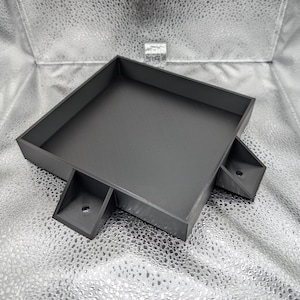 May include: A square, black tray with raised edges and two angled support structures. The tray has a textured surface and a small hole in each support. The item is likely 3D printed.