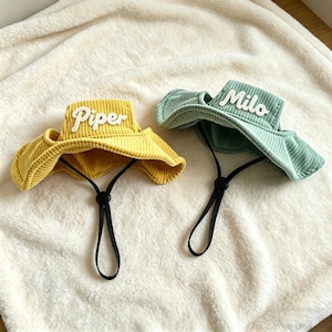 May include: Two corduroy bucket hats with chin straps and toggles. One hat is yellow with the white embroidered name Piper, and the other is light green with the white embroidered name Milo. Both hats feature a textured ribbed fabric finish.