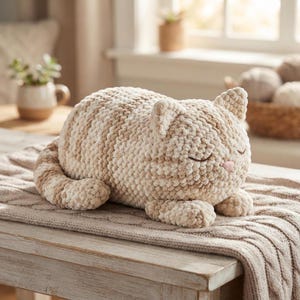 Puede incluir: Un juguete de peluche con forma de gato hecho de hilo grueso y suave con rayas alternas en color crema y bronceado claro. El gato está en posición de descanso con los ojos cerrados, una pequeña nariz rosa bordada y una cola larga.
