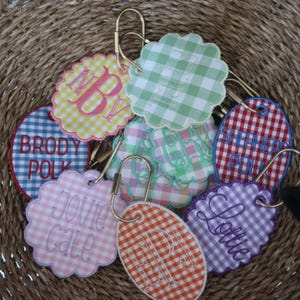 May include: A collection of embroidered gingham fabric keychains in various colors, including yellow, green, pink, orange, red, and purple. Each keychain features a unique monogram or name in cursive script, attached to a gold-colored clasp.