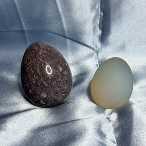 May include: Two polished stone eggs. One is a dark purple color with a speckled texture. The other is a pale, translucent yellow. Both eggs are smooth and oval-shaped, resting on a blue satin surface.