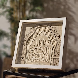 Op de afbeelding: Een ingelijst kunstwerk in beige, met ingewikkelde lasergesneden Arabische kalligrafie. Het witte frame omringt het gedetailleerde ontwerp, met geometrische patronen en schrift. Het kunstwerk wordt op een donkere ondergrond getoond.