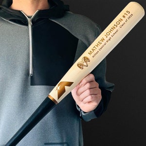 Könnte beinhalten: Ein Baseballschläger aus Holz mit einem hellen Naturholz-Lauf und einem schwarzen Griff. Der Lauf zeigt ein goldenes Widder-Logo, ein goldenes Sternsymbol und den Text MATHEW JOHNSON #15 Hilton Central High School - Class of 2026 in goldener Schrift.