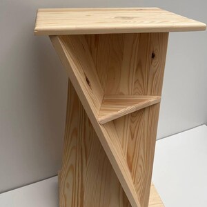 May include: A light-colored wooden side table with a geometric design. The table features a square top, a triangular shelf, and a base with small wheels. The wood grain is visible throughout the structure.