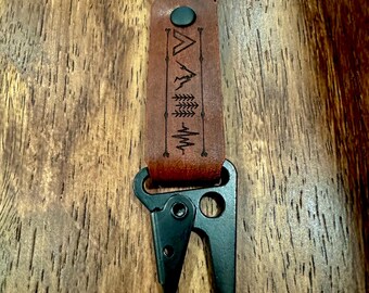 May include: A brown leather keychain with a black metal clasp and ring. The leather strap features a mountain and tree design with a heartbeat line. The keychain is designed for outdoor enthusiasts.