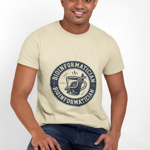 Op de afbeelding: Beige T-shirt met een cirkelvormige afbeelding met het woord "BIOINFORMATICIAN" rond een koffiekopje en een DNA-streng. Het shirt wordt gedragen door een persoon in een spijkerbroek.