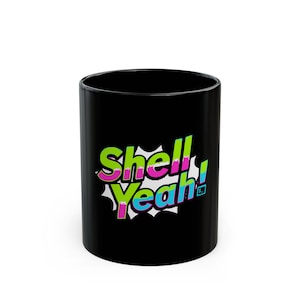Op de afbeelding: Een zwarte keramische mok met de tekst "Shell Yeah!" in een kleurrijke stripboekstijl. De tekst is in groen, roze en blauw, met een witte burst-achtergrond. De mok is cilindrisch en heeft een glanzende afwerking.