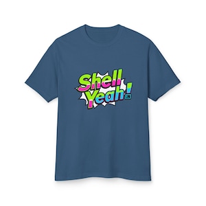 Op de afbeelding: Een marineblauw t-shirt met de tekst "Shell Yeah!" in een kleurrijk stripboekstijl lettertype. De tekst is zwart omlijnd en geplaatst tegen een witte burst-achtergrond. Het shirt heeft korte mouwen en een ronde hals.