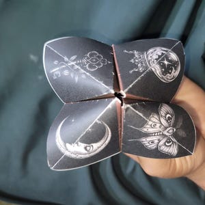 May include: A black paper fortune teller featuring white line art illustrations on each of its four flaps. The designs include a detailed ornate key, a glowing crystal ball, a crescent moon with a face, and a moth with intricate wing patterns.