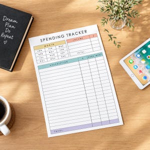 May include: A white paper spending tracker sheet with black text and colored headers in yellow, peach, teal, and purple. The form includes sections for month, income, and a detailed list with columns for description, dollar amount, need, and want.