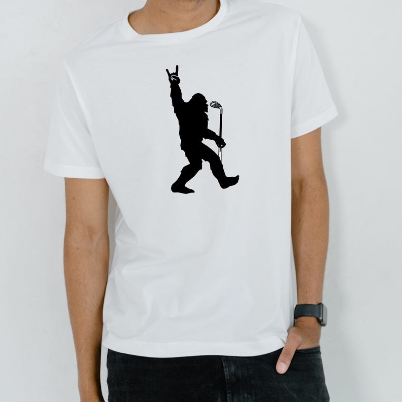 May include: White t-shirt featuring a black silhouette of Bigfoot holding a golf club and making a rock-and-roll hand gesture. The shirt is a classic crew neck style.