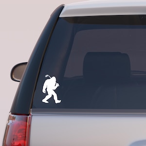 May include: White vinyl decal of Bigfoot walking and carrying a golf club. The decal is applied to the rear window of a vehicle. The design is a silhouette of the legendary creature.