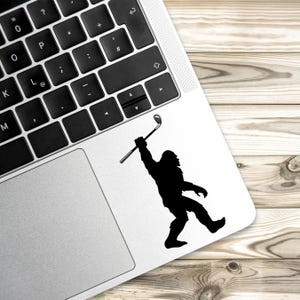 May include: Black silhouette sticker of Bigfoot swinging a golf club. The sticker is placed on a laptop next to the keyboard. The background is a light brown wooden surface.