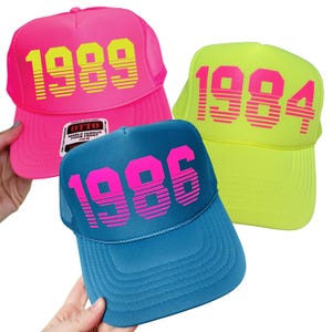 May include: Three foam trucker hats featuring horizontal striped numbers on the front. One is hot pink with yellow 1989 text, one is neon yellow with pink 1984 text, and one is teal with pink 1986 text. Each hat has a mesh back and a structured brim.