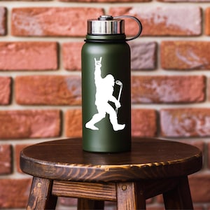 May include: A dark green insulated water bottle with a silver lid and black handle. A white graphic of Bigfoot holding a golf club and making a rock-and-roll hand gesture is on the front. The bottle is sitting on a wooden stool.