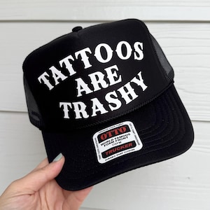 May include: Black trucker hat with a white mesh back. The front of the hat has white text that reads "TATTOOS ARE TRASHY". The hat has a black label with white text that reads "OTTO WORLD FAMOUS FOAM FRONT OSFM TRUCKER DURABLE SNAP CLOSURE".
