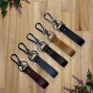 May include: Five leather keychains in various colors: black, brown, tan, dark gray, and burgundy. Each keychain features a silver-colored clasp and a leather strap. The keychains are arranged on a wooden surface with decorative greenery.