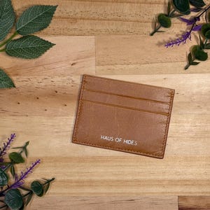 May include: A brown leather card holder with three card slots, the text "HAUS OF HIDES" is printed on the front. The card holder is on a wooden surface, with green and purple floral accents.