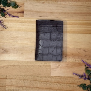 May include: A dark grey, rectangular wallet with a crocodile skin texture. The wallet is centred on a light brown wooden surface, with sprigs of greenery and purple flowers in the corners.