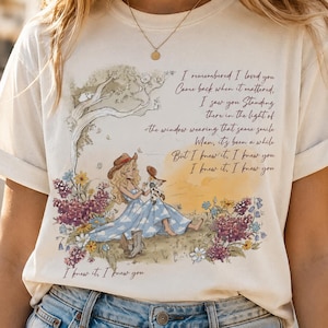 Op de afbeelding: Een crèmekleurig t-shirt met een gedrukte illustratie van een persoon met een cowboyhoed en een blauwe jurk, zittend in een veld met paarse en gele bloemen. De afbeelding bevat de tekst: I remembered I loved you, Come back when it mattered, I saw you standing there in the light of the window wearing that same smile, Man, it's been a while, But I knew it, I knew you, I knew it, I knew you.