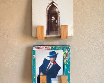 May include: Two vinyl record albums displayed on a wall with wooden holders. The top album cover features a man in a doorway, and the bottom album cover features a man in a blue suit and hat. The wall is painted a light beige color.