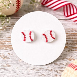 May include: Baseball-themed stud earrings. Each earring is a small, white circle with red stitching, mimicking the design of a baseball. The earrings are displayed on a white surface, with a baseball and red and white ribbon in the background.