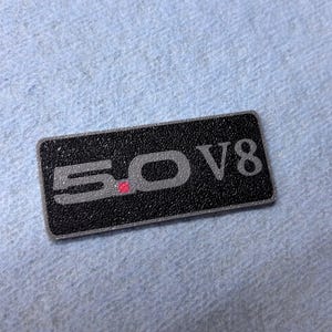 May include: Black rectangular emblem with the text "5.0 V8" in silver. The emblem has a textured surface and a silver border. The "0" in "5.0" has a small red dot.
