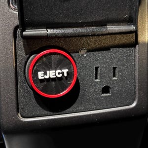May include: Close-up of a black car console featuring a power outlet and an "EJECT" button with a red border. The outlet has two vertical slots and a circular bottom slot.