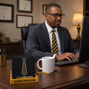 Könnte beinhalten: Ein Satz schwarzer quadratischer Untersetzer mit einem gelben Krawattenmuster und dem Text "ALPHA PHI ALPHA". Die Untersetzer befinden sich in einem gelben Halter. Ein weißer Becher steht auf einem passenden Untersetzer. Eine Person im Anzug arbeitet an einem Computer.