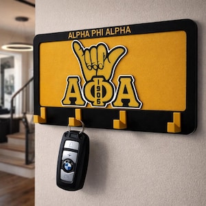 Peut inclure: Porte-clés noir avec des accents jaunes et le texte "ALPHA PHI ALPHA". Le porte-clés présente un fond jaune avec le symbole de la main et les lettres de la fraternité. Une clé de voiture est suspendue à l'un des crochets.